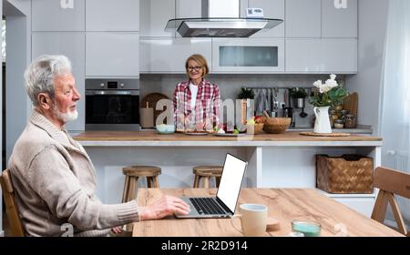 Happy senior mature man husband remote working or distance learning online from home office using laptop computer while her older wife cooking breakfa Stock Photo