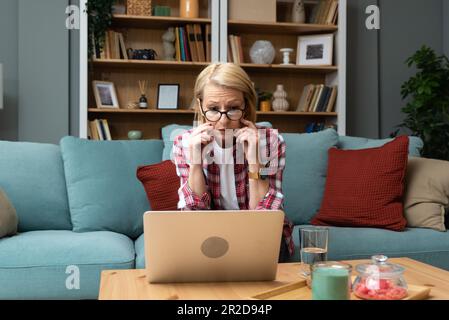 Worried middle-aged 70s lady sit on sofa in living room read negative unexpected news on laptop, anxious mature woman rest on couch feel distressed wi Stock Photo