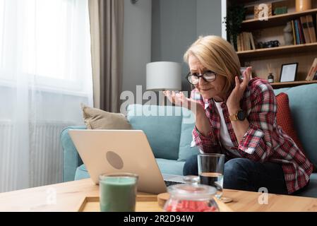 Worried middle-aged 70s lady sit on sofa in living room read negative unexpected news on laptop, anxious mature woman rest on couch feel distressed wi Stock Photo