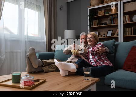 Laughing aged couple, man and woman watching tv, comedy show or movie and eating popcorn snack, sitting on cozy couch at home, mature family, man and Stock Photo