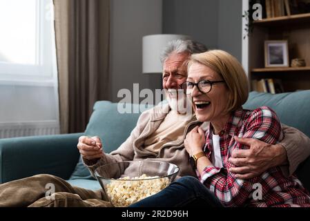 Laughing aged couple, man and woman watching tv, comedy show or movie and eating popcorn snack, sitting on cozy couch at home, mature family, man and Stock Photo