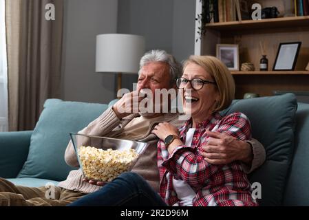 Laughing aged couple, man and woman watching tv, comedy show or movie and eating popcorn snack, sitting on cozy couch at home, mature family, man and Stock Photo