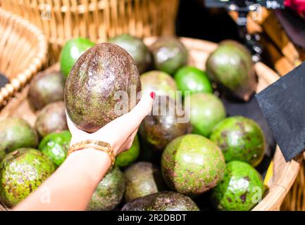 The woman's right-hand holding and select buy big  avocado color brown and green in the basket in the department store, the photograph is close up, bl Stock Photo