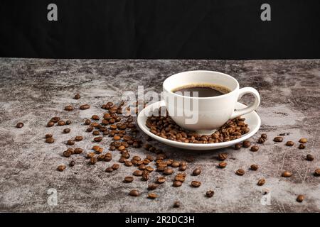 A close-up of hot coffee in a white cup is placed on a cement floor table, lots of roasted coffee beans are in the coffee cup saucer, and around, smok Stock Photo