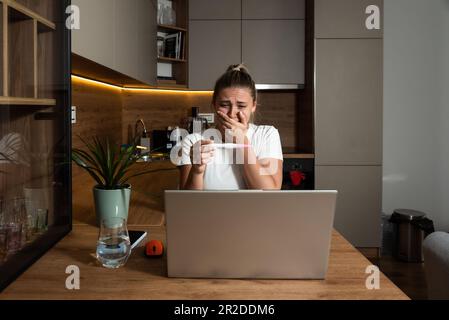 Young woman surprised negative facial reactions on positive result on pregnancy test. Young girl talking on video call on laptop computer with her mot Stock Photo