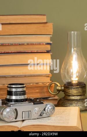 An open old book on which lies an analog photo camera. In the background, a pile of old books and a lamp stylized as an antique kerosene lamp. Items s Stock Photo