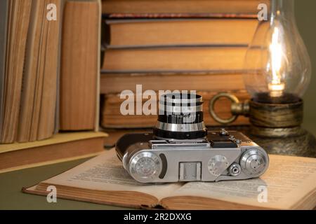 An open old book on which lies an analog photo camera. In the background, a pile of old books and a lamp stylized as an antique kerosene lamp. Items s Stock Photo