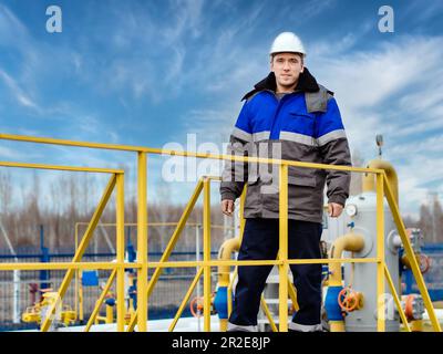 Portrait of worker in white helmet and winter pea jacket at industrial gas facility. Middle-aged Caucasian man stands in full growth on territory of gas distribution station. Genuine worker. Stock Photo