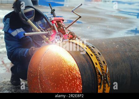 Welder in mask cuts large-diameter pipe with autogenous gun and sparks fly. Device for cutting metal pipes. Cutting metal with oxygen. Authentic outdoor workflow. Stock Photo