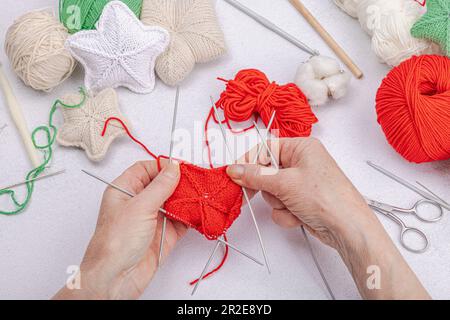 Women's hands knit New Year's decor with knitting needles. Soft voluminous stars, skeins of threads, accessories. Hobby, handmade concept. Light stone Stock Photo