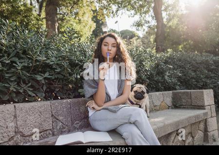 Focused young and curly woman in casual clothes holding marker and looking at camera near notebook with copy space and pug dog sitting on stone bench Stock Photo