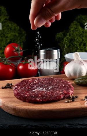 Fresh raw burger cutlet with spices and herbs on a black background. man salts a raw burger patty. Stock Photo