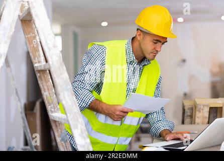 Engineer with drawings in his hands, checks the house reconstruction plan on a laptop Stock Photo