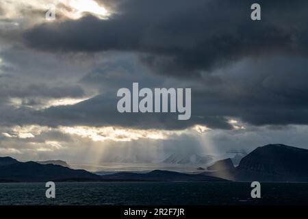 Landscape photo of the sun bursting through openings in the clouds to shine on a barren, snowy scene, Ny-Ålesund, Spitsbergen, Norway, Europe Stock Photo