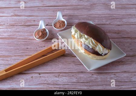 Delicious chocolate-covered cream eclair and white ceramic spoons with chocolate and cocoa on the side with wooden tongs Stock Photo