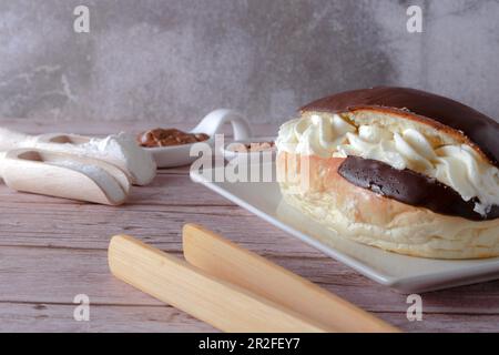 Delicious chocolate-covered cream eclair and white ceramic spoons with chocolate and cocoa on the side with wooden tongs Stock Photo
