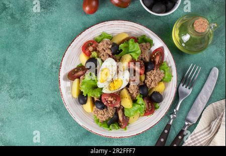 Salad with canned tuna, boiled potatoes, cherry tomatoes, eggs, olives and lettuce, dressed with olive oil on green background, Top view Stock Photo