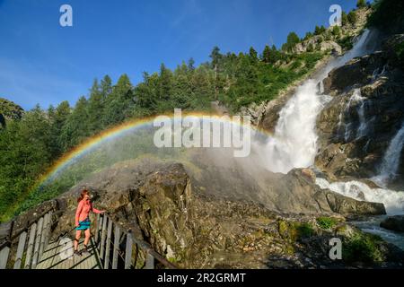 Woman hiking standing on bridge looking at Rutor waterfalls with rainbow, Rutor falls, Rutor group, Graian Alps, Aosta, Italy Stock Photo