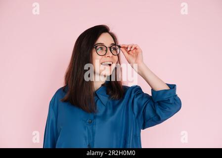 Smiling woman touching glasses, pondering and looking for ideas in empty space: Isolated portrait on pink background Stock Photo