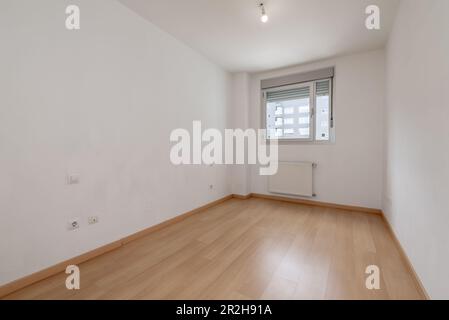 An empty room with white painted walls, a light wooden floor, a single aluminum window with a view and cheap aluminum radiators Stock Photo