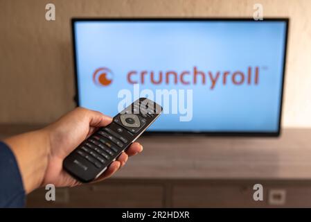 Brazil, Rio de Janeiro - May 19, 2023: In this photo illustration a person holds the TV remote control and in the background, the Crunchyroll logo. Stock Photo