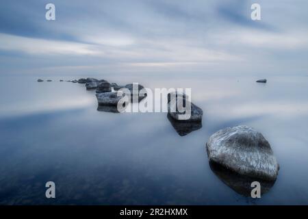 Stones in the water lie in a slight arc. Smooth water surface. Clouds are reflected in the water. Kronvalds Fiskeläge, Klintehman, Gotland County, Swe Stock Photo