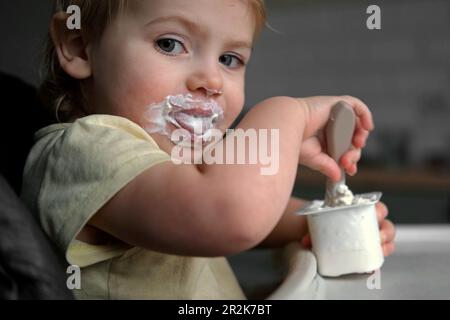 Young Kid Eating Blend Mashed Feed Sitting in High Chair. Baby Weaning. Little Girl Learning to Eat Yogurt, Feeding Himself. Small Hand with Spoon. Br Stock Photo