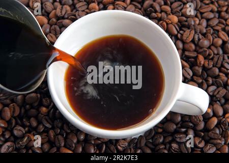 Pouring black coffee into white ceramic cup. Top view. Close up. Hot coffee drink pour into cup from shiny moka pot coffee maker. Roasted coffee beans Stock Photo