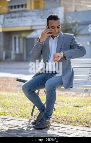 Young latin man sitting on a square bench talking on mobile phone and looking at his watch. Stock Photo