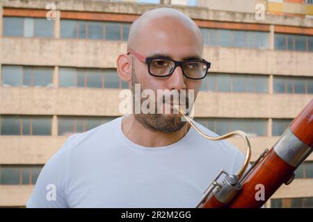 closeup portrait of young latino man with glasses, beard and bald, wearing white t-shirt, standing playing bassoon on apartment balcony with urban bui Stock Photo