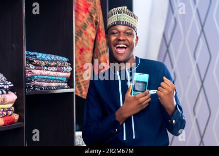 african man wearing african attire selling in a fashion market using a point on sale machine celebrating successful transaction. Payment method, cashl Stock Photo