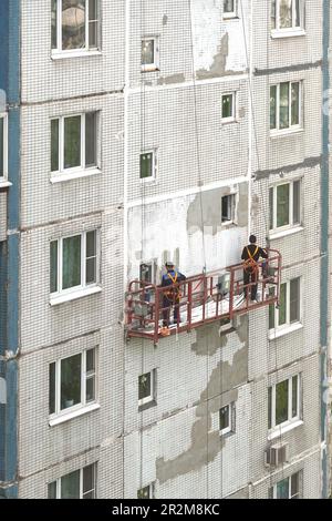 Two workers in construction cradle on metal cables repairing wall surface on block of flats house on the high Stock Photo