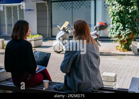 Back view shot of two businesswomen sitting on a bench in the city and having a conversation. Brunette girl holding a lap top on her lap Stock Photo