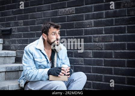 Young sad sick poor homeless man sitting on the street need help feeling lonely and depressed because he is abandoned from family and friends when he Stock Photo