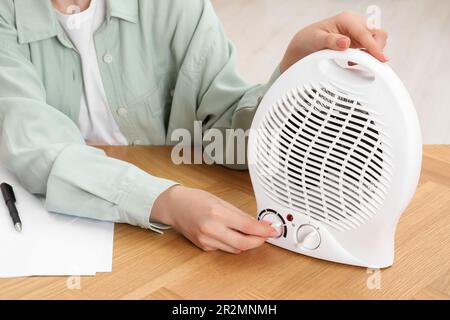 Young woman adjusting temperature on modern electric fan heater at wooden table, closeup Stock Photo