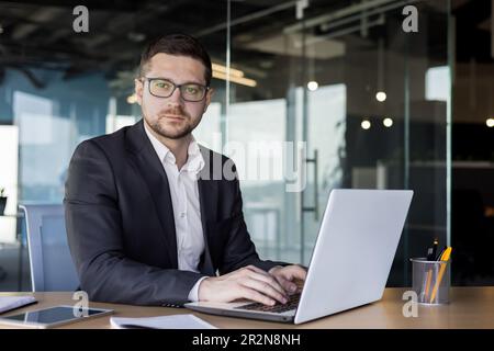 Portrait of a young man, a businessman, a lawyer, who works in an office at a desk and concentrates on a laptop. He looks confidently and seriously in Stock Photo