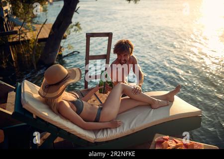 A young couple in swimsuit enjoying the sun and drink on the river bank on a beautiful summer day. Summer, river, vacation Stock Photo