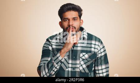 Shh be quiet please. Young indian man presses index finger to lips makes silence gesture sign do not tells secret, stop gossip. Handsome bearded hindu guy isolated alone on beige studio background Stock Photo