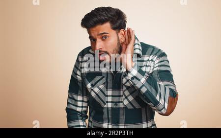I cant hear you. What. Young indian man trying hear, looking confused, frowning, keeping arm near ear for louder voice, asking to repeat, to hear information, deafness. Hindu guy on beige background Stock Photo