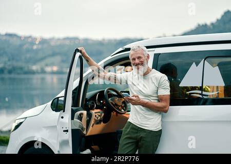 Happy older man traveler standing near camper van using mobile phone. Stock Photo