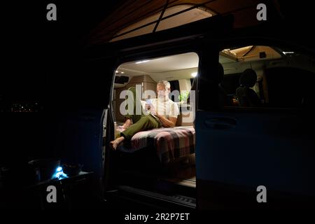 Old man traveler sitting in camper van using mobile in camping at night. Stock Photo