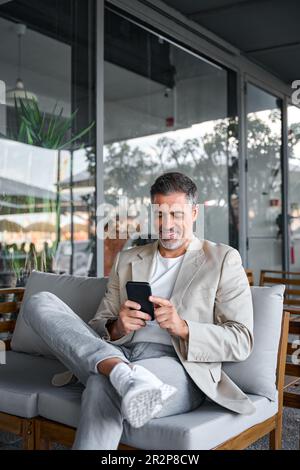 Happy relaxed mid aged business man sitting in outdoor cafe using smartphone. Stock Photo