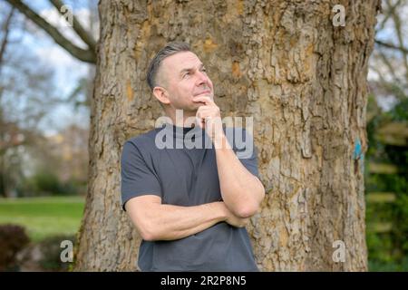 Thoughtful grey-haired man standing in front of a tree in the park and looking up Stock Photo