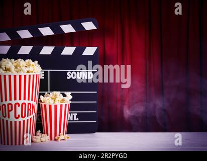 Delicious popcorn and clapperboard on wooden table against closed red main curtain, space for text Stock Photo