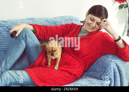 Attractive smiling woman lying on the sofa with her dog sitting on her in the living room of her house Stock Photo