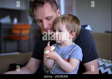 Little toddler eating banana held by his father Stock Photo