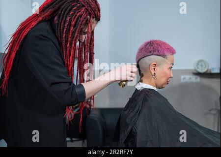 The hairdresser shaves the temple of a female client. Asian woman with short pink hair in barbershop. Stock Photo