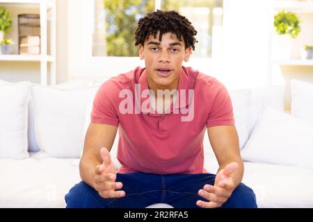Portrait of biracial young man with dreadlocks gesturing and looking at camera while sitting on sofa Stock Photo