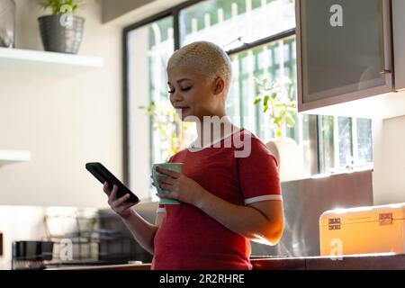 Happy biracial woman using smartphone and drinking coffee in kitchen. Lifestyle, communication and domestic life. Stock Photo