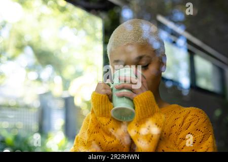 Happy fashionable unaltered biracial woman enjoying coffee at home, seen through window. Relaxation, free time and domestic lifestyle. Stock Photo